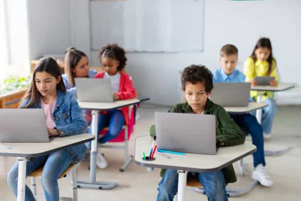 Diverse school children sitting at desks with laptops, studying using digital gadgets. Modern elementary education concept