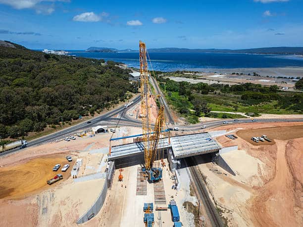 Aerial view of a large construction project featuring a road bridge over a railroad track. Workers and vehicles are evident, surrounded by urban planning efforts. The site is located near the sea, showcasing natural landscapes and water. A modern development representing engineering progress.